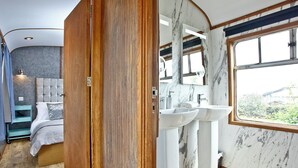 A bedroom with a bed, a nightstand, a dresser, and a bathroom with two sinks and a large window. - Brunel Boutique Railway Carriage 3 (Dawlish Warren)