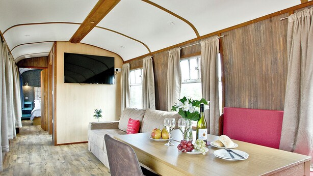 A dining area with a wooden table, chairs, a sofa, and a TV mounted on the wall. - Brunel Boutique Railway Carriage 3 (Dawlish Warren)