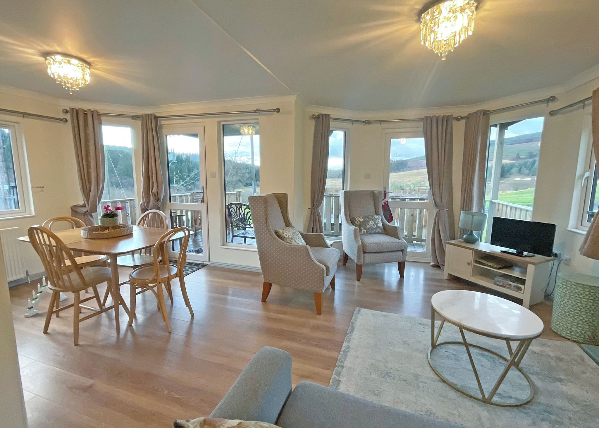 A spacious living area with a dining table, chairs, sofas, a TV, and large windows with curtains.