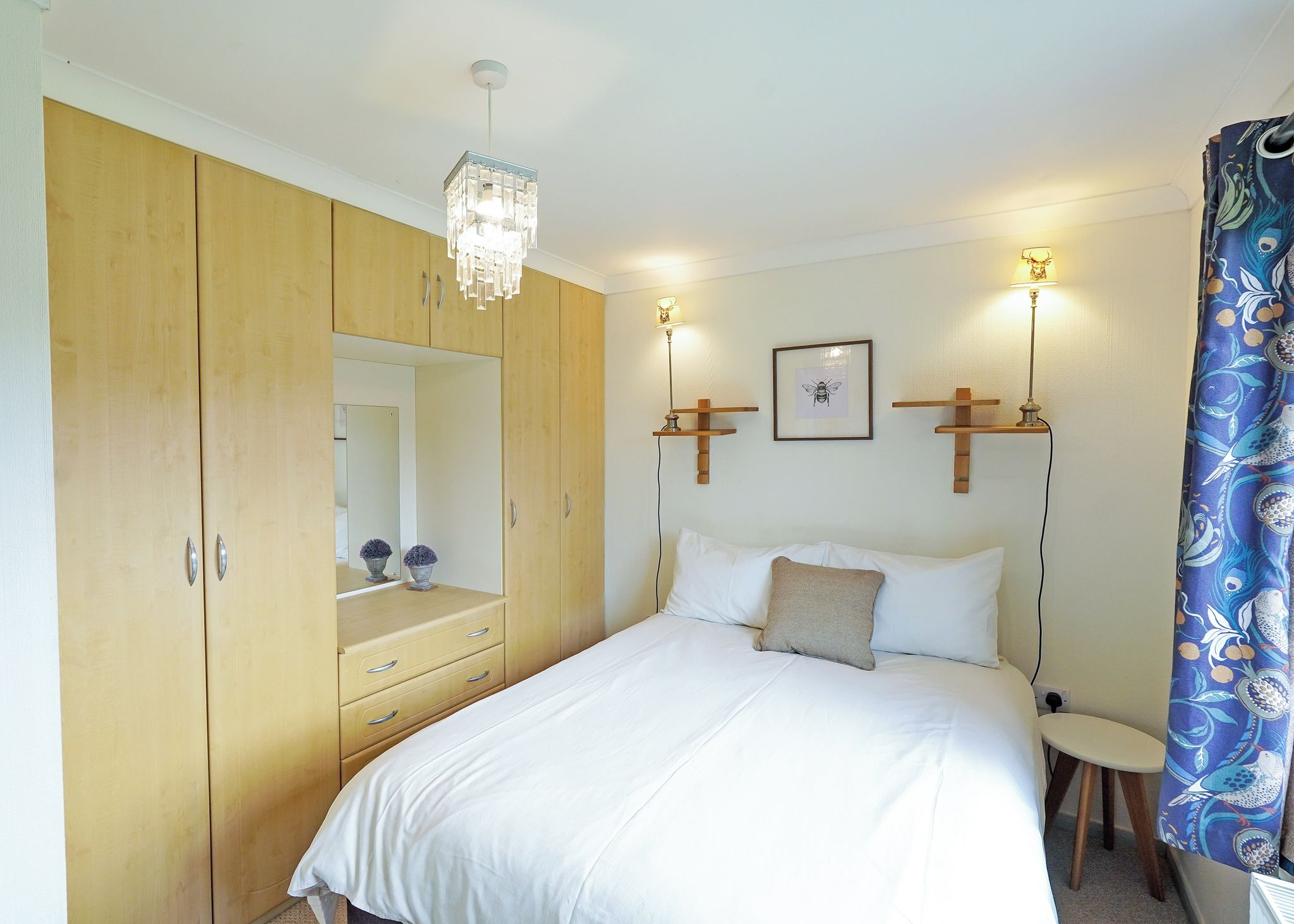 A bedroom with a bed, a wooden wardrobe, a mirror, a bedside table, and wall-mounted lights.