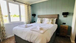 Cama bem arrumada com lençóis e toalhas brancas, mesa de cabeceira com abajur e janela com cortinas.