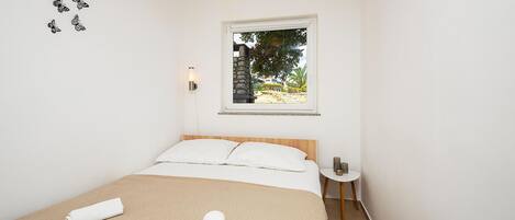A bedroom with a bed, a window with a view of greenery, a wall-mounted light, and a small table with a vase.