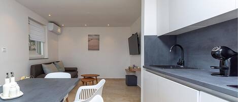 Una cocina moderna con encimera oscura, armarios blancos y grifo negro. Hay un comedor con una mesa y sillas, y un sofá al fondo.