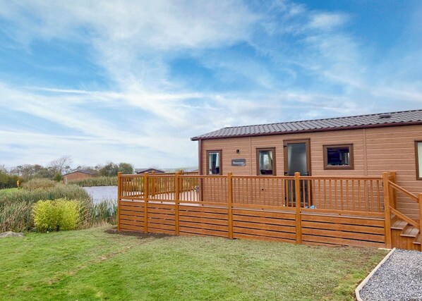 Une cabane en bois avec une terrasse, une pelouse et un plan d’eau en arrière-plan.