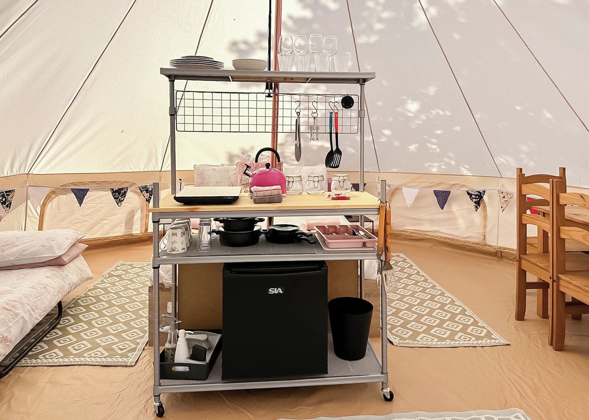 A tent interior with a kitchen cart, a bed, a chair, and a table with a tent wall in the background.