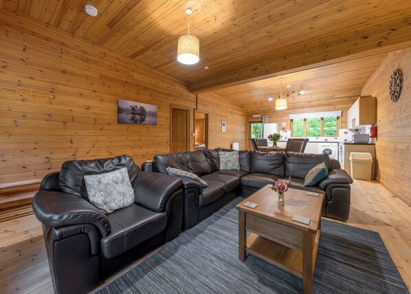 A wooden interior with a sectional sofa, a wooden coffee table, and a kitchen area visible through an open door.
