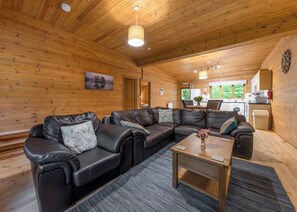 A wooden interior with a sectional sofa, a wooden coffee table, and a kitchen area visible through an open door. - Woodland Rowan Lodge (Ellesmere)