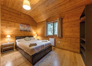 A wooden cabin bedroom with a bed, two bedside lamps, a radiator, a window with curtains, and a painting on the wall. - Woodland Rowan Lodge (Ellesmere)