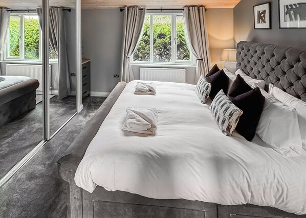 Habitación de hotel con una cama grande, cabecera gris y vistas a zonas verdes a través de las ventanas.