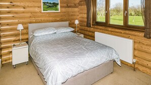 A log cabin bedroom with a bed, nightstands, a lamp, a window, and a radiator. - One double with en suite shower and one twin. (Laxfield, Nr Woodbridge)