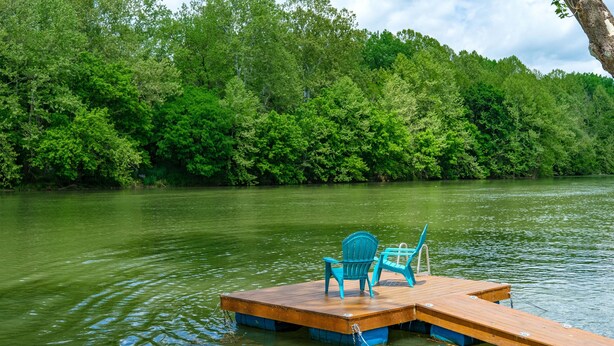 A wooden dock with two chairs and a small table, surrounded by calm water and dense green foliage. - RIVER FRONT HOME AMAZING VEIWS AND HOT TUB (Shenandoah)