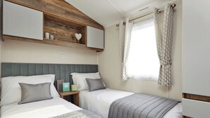 Un dormitorio con dos camas, cabeceros de madera, una ventana con cortinas y una mesita de noche con lámpara.
