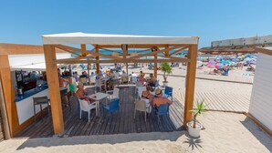 Een terras buiten met houten tafels en stoelen, met uitzicht op een strand met parasols en mensen.