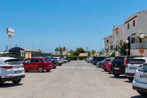 Ein Parkplatz mit mehreren in Reihen geparkten Autos, ein Gebäude mit der Aufschrift 'MERCADONA' und einem strahlend blauen Himmel.
