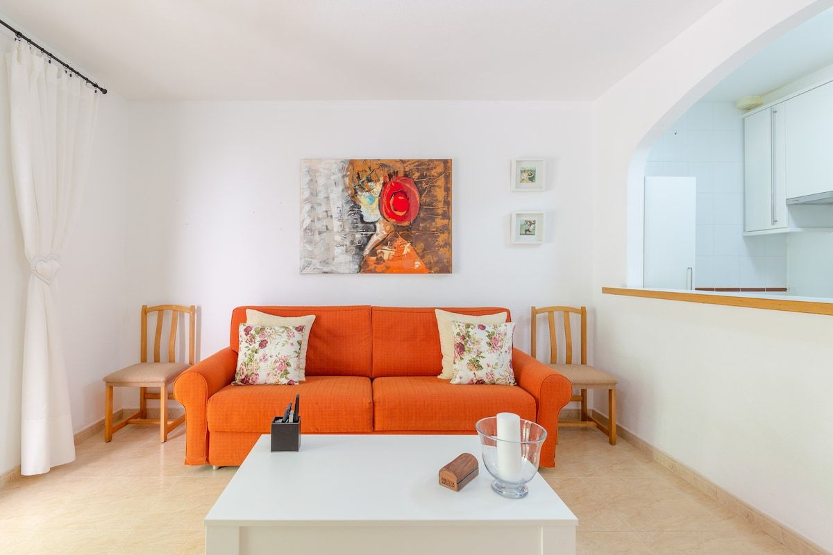 A living room with an orange sofa, two chairs, a white coffee table, and a painting on the wall.
