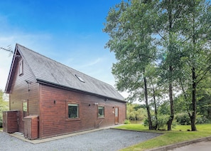 Un bâtiment en bois avec un toit en pente, une allée gravillonnée et une zone herbeuse avec des arbres.