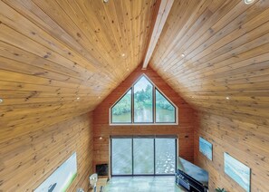 A wooden interior with a sloped ceiling, large windows, and a view of greenery outside.