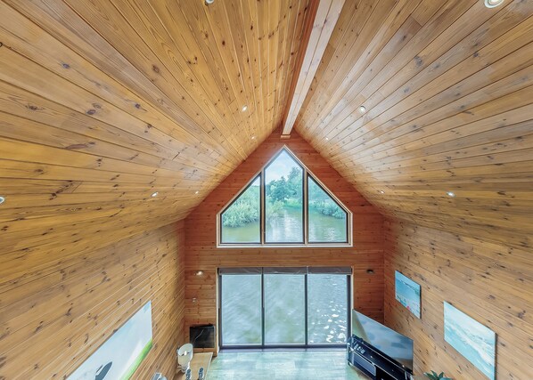 A wooden interior with a sloped ceiling, large windows, and a view of greenery outside. - Lower Lake Boathouse (Anglesey)