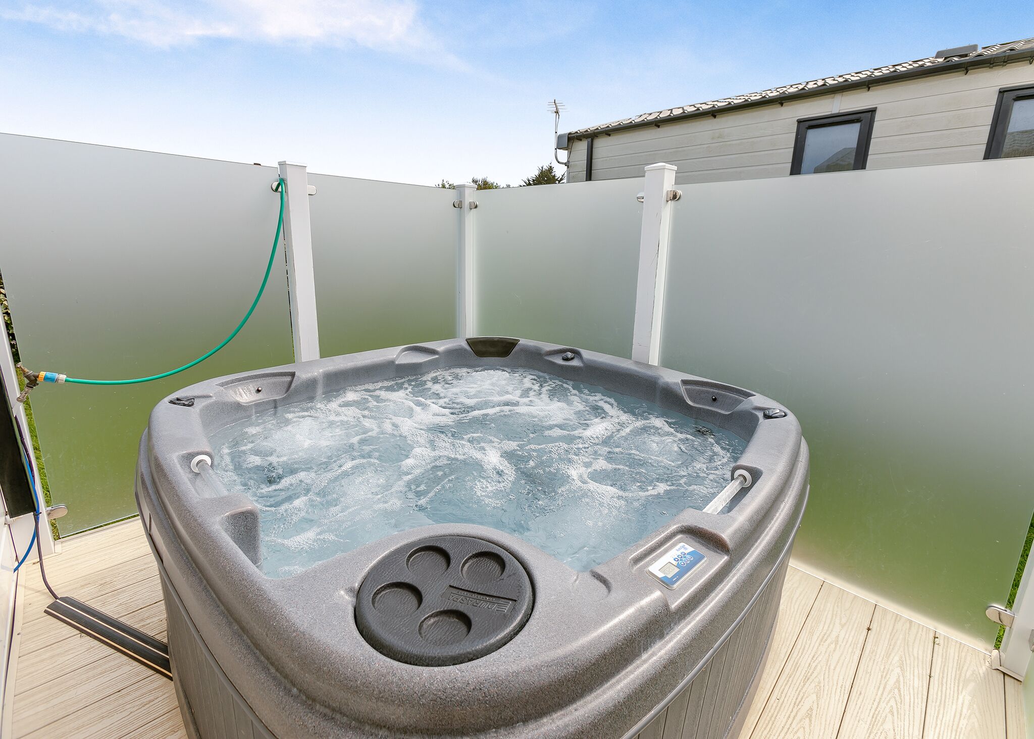 Un jacuzzi con panel de control, rodeado por una terraza de madera y una valla verde.
