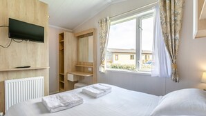 A hotel room with a bed, a TV mounted on the wall, a window with curtains, and a wooden wardrobe. - Luxurious holiday home. (Looe)