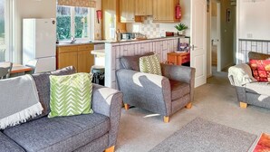 A compact living space with a kitchenette, two grey sofas with patterned cushions, a dining area, and a view of the outdoors. - Country Lodge Six Spa (Tintagel)