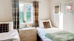 A bedroom with two beds, a window with curtains, a bedside table with a lamp, a small plant, and a wall with framed pictures. - Country Lodge Six Spa (Tintagel)