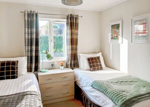 A bedroom with two beds, a window with curtains, a bedside table with a lamp, a small plant, and a wall with framed pictures. - Country Lodge Six Spa (Tintagel)
