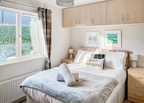 A bedroom with a bed, a window with curtains, a bedside table with a lamp, and a wall with framed pictures.
