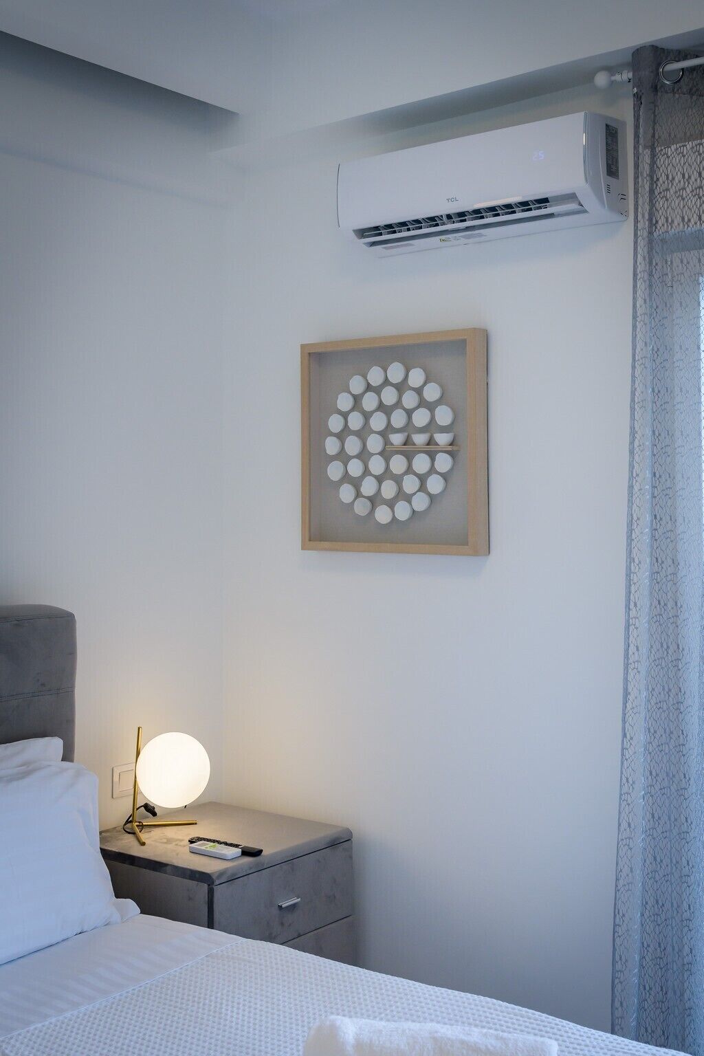 A hotel room with a bed, a bedside table with a lamp, a framed abstract artwork, and an air conditioning unit.