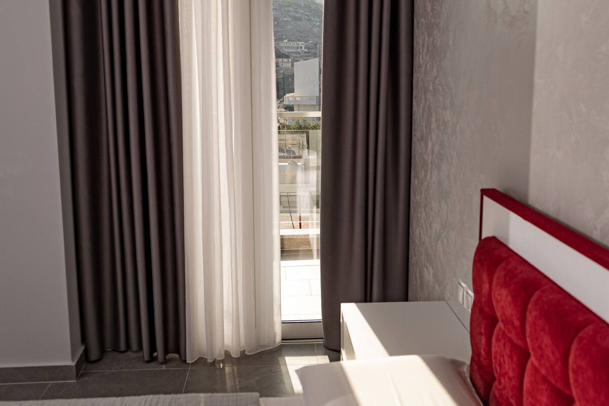 A hotel room with a bed, a red headboard, a window with curtains, and a view of a cityscape.