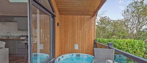 A modern outdoor hot tub area with glass doors leading to a kitchen, a wooden deck, and a glass table with a clear view of the sky.