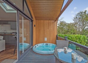A modern outdoor hot tub area with glass doors leading to a kitchen, a wooden deck, and a glass table with a clear view of the sky.