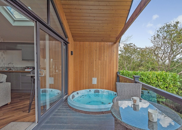 A modern outdoor hot tub area with glass doors leading to a kitchen, a wooden deck, and a glass table with a clear view of the sky.