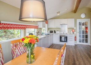 A kitchen with white cabinets, a dining table with a vase of flowers, a window with a view of greenery, and a clock on the wall. - Very comfortable lodge with one double and one twin. (Millom)