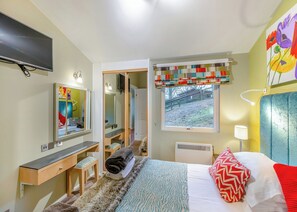 Un dormitorio con cama, televisor, escritorio, espejo, ventana con vistas y una obra de arte colorida en la pared.