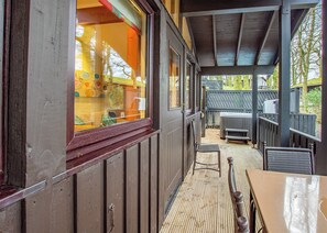 A covered outdoor seating area with wooden benches and a table, a wooden deck, and a view of a building with colorful windows.