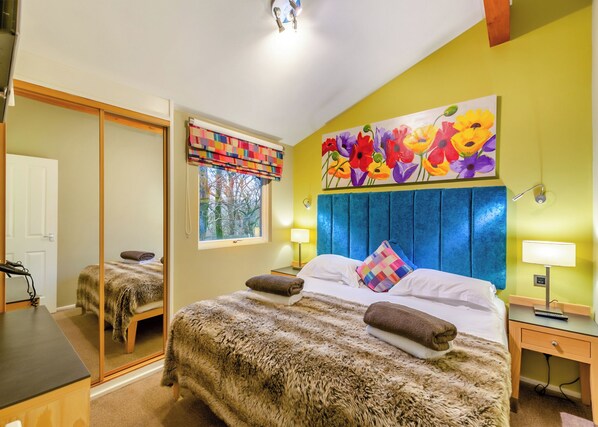 Une chambre avec un grand lit, une fenêtre agrémentée d’une lambrequin colorée, un tableau de fleurs accroché au mur et un miroir dans la pièce adjacente.