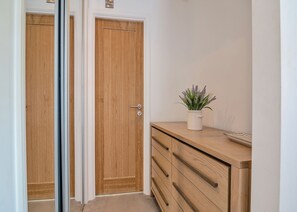 A hallway with a wooden door, a white wall, and a wooden cabinet with a plant on top.