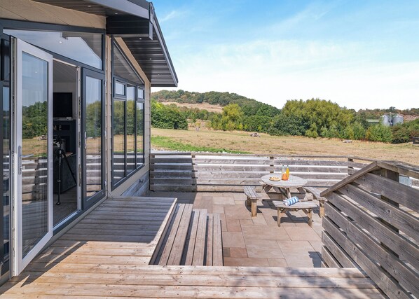 A modern outdoor deck with a glass door leading to a patio area, a wooden table with a bottle and a plate, and a view of a hilly landscape. - 2 bedroom accommodation in Cannington (Cannington)