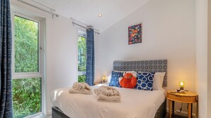 Ein Schlafzimmer mit einem großen Bett, einem Nachttisch mit Lampe, einem Fenster mit Vorhängen und einem Bild an der Wand.
