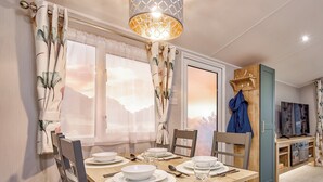 A dining area with a table set for four, a TV, and a wooden cabinet with a coat hanging on it. - Signature 3 Bed Caravan Pet (Morpeth)