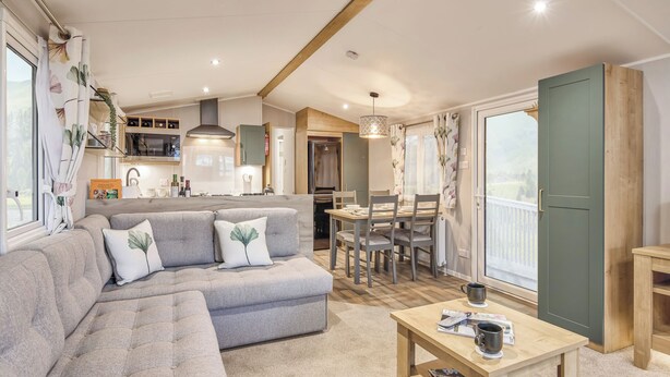 A modern living room with a sectional sofa, a dining table, and a kitchen in the background. - Signature 3 Bed Caravan Pet (Morpeth)
