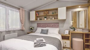 A bedroom with a bed, bedside tables, a vanity with a mirror, and built-in shelving with books and decorative items. - Signature 3 Bed Caravan Pet (Morpeth)