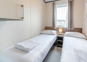 A hotel room with two single beds, a bedside table with a lamp, a window with curtains, and a wall-mounted shelf.