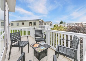 A balcony with two chairs and a small table, a white railing, and a view of mobile homes in the background.