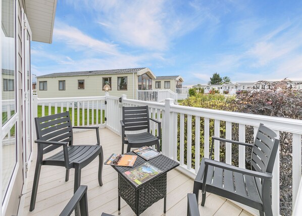 A balcony with two chairs and a small table, a white railing, and a view of mobile homes in the background. - Luxurious 14ft-wide holiday home. (Looe)