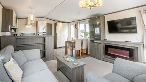 A modern living room with grey sofas, a coffee table, a fireplace, a TV, and a kitchen area with cabinets and a microwave. - Luxurious 14ft-wide holiday home. (Looe)