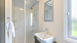 A modern bathroom with a glass shower enclosure, a white sink, and a mirror above it. - Luxurious 14ft-wide holiday home. (Looe)