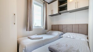 A room with two beds, a window with curtains, and built-in cabinets above the beds. - Luxurious 14ft-wide holiday home. (Looe)