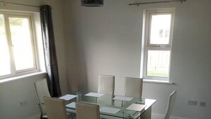 Un coin repas avec une table en verre et six chaises, deux fenêtres avec des rideaux et un luminaire suspendu.